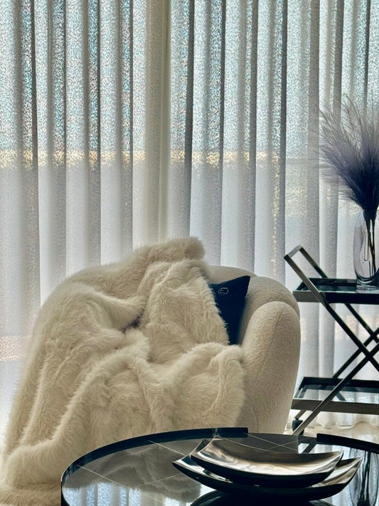 Plaid en fausse fourrure crème moelleux posé sur fauteuil beige dans salon moderne avec rideaux blancs